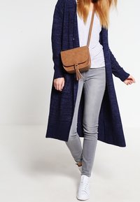 Cárdigan largo navy, camiseta gris clara, jeans gris claro, bolso cruzado marrón con flecos y zapatillas blancas. Texturas suaves, ajuste casual.