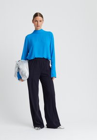 Langärmliges, hochgeschlossenes Oberteil in leuchtendem Blau, kombiniert mit schwarzen, weit geschnittenen Hosen. Das Model hält eine silberne Tasche und trägt metallische Schuhe.