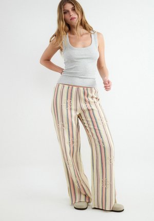 Žena oblečená v sivom tielku bez rukávov a širokých pásikavých nohaviciach s vysokým pásom a vyšívanými vzormi, stojaca pred bielym pozadím.