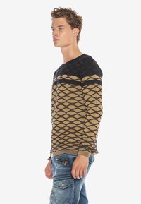 Braun und schwarz gemusterter Pullover mit einem Diamantenmuster, mit Rundhalsausschnitt und gerippten Bündchen. Getragen mit blauen Cargo-Hosen.