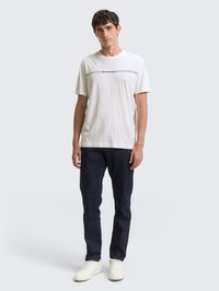 Λευκό βαμβακερό μπλουζάκι με μαύρη οριζόντια εκτύπωση λογότυπου, συνδυασμένο με σκούρο μπλε τζιν και λευκά αθλητικά παπούτσια. Απλός, καθημερινός σχεδιασμός outfit.