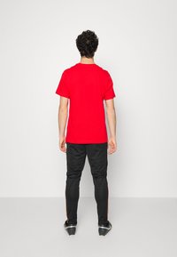 Muž stojící zády, oblečený v jasně červeném tričku s krátkým rukávem, černých sportovních kalhotách s oranžovými pruhy a černých kopačkách na bílém pozadí.