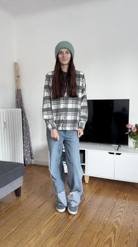 Hall kastanjas ja valge ruudulise mustriga nupp-särk, sinised laiad teksad friteeritud äärtega, hall kudumine müts, mustrilised spordijalatsid; puidust põrand ja teler taustal.