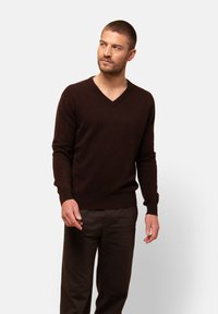 Ciemnobrązowy sweter w serek wykonany z miękkiego, strukturalnego materiału, z długimi rękawami i prążkowanymi mankietami, zestawiony z dopasowanymi brązowymi spodniami.