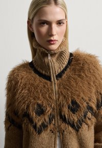 Brauner gestrickter Pullover mit einem Kragen aus Kunstfell, der schwarze geometrische Muster aufweist und einen Reißverschluss hat. Weicher Stoff und hochgeschlossenes Design.
