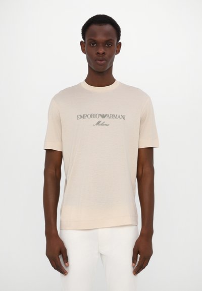 Tricou scurt bej cu mâneci scurte, dintr-un material moale, cu decolteu rotund și logo-ul "Emporio Armani Milano" în verde închis. Design simplu.