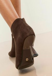 Botine din suede maro, cu toc înalt, având o curbă netedă, vârful închis și un detaliu cu fermoar pe spate. Tocul are un finisaj lucios.