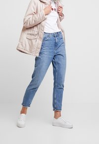 Ljusbeige vadderad jacka över en vit t-shirt med ett blommönster, kombinerat med blå högmidjade jeans och vita sneakers.