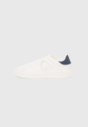 White low-top sneaker with navy heel tab, lace-up closure, and embossed shield logo on side, shown in right profile on white background.