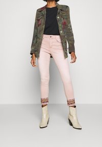 Kurtka w kamuflażu z dwoma przednimi kieszeniami; czarna koszulka; różowe dżinsy z haftowanymi akcentami na hemie; beżowe botki na obcasie.