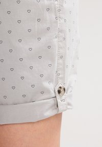 Shorts gris clair avec petits motifs de cœurs noirs, ourlet roulé et détail de bouton marron sur la couture latérale.