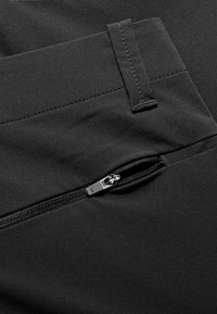 Close-up van een zwart stof met een gedeeltelijk open ritssluiting en een gestikte riemlus op een kledingstuk.