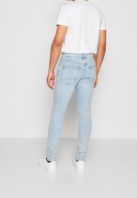 Hombre vestido con jeans skinny de color azul claro, camiseta blanca y zapatillas blancas, de pie contra un fondo blanco liso, mirando hacia atrás.