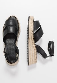 Černé kožené sandály na klínovém podpatku s pletenými jutu platformami a páskami kolem kotníku, jedny sandály zobrazeny zboku a druhé shora.