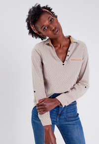 Blusa beige con estampado, de escote en V, mangas largas y detalles de botones. Lleva puesta con jeans azules, cuenta con un bolsillo en el pecho y cuello.