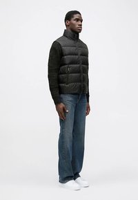 Czarna pikowana kamizelka z wzorzystą tkaniną i wysokim kołnierzem, w połączeniu z niebieskimi prostymi dżinsami i białymi tenisówkami. Postawa stojąca na jednolitym tle.