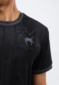 Κοντινό πλάνο ενός ατόμου που φοράει μαύρο κοντομάνικο πουκάμισο με διακριτικό σχέδιο από φίδι και μικρό ασημένιο λογότυπο με δόντι φιδιού στο στήθος.