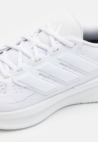 Gros plan d'une chaussure de sport blanche avec tissu texturé, trois bandes en plastique, lacets blancs et semelle nervurée.