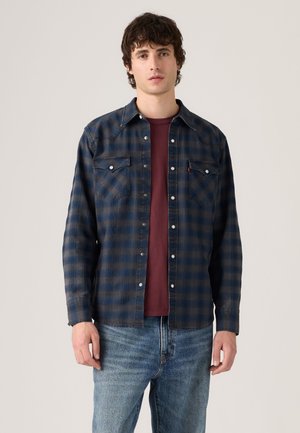 Jeune homme portant une chemise à carreaux bleue et noire boutonnée sur un t-shirt bordeaux et un jean bleu clair, debout devant un fond uni.