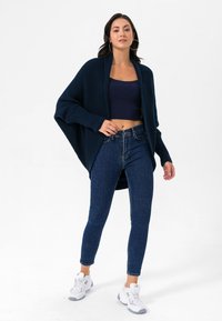 Tumedast ribadega kardigan tume lühikese topi peal, koos tumedate siniste kitsaste teksade ja valgete sportlike jalatsitega. Naine seisab valgel taustal.