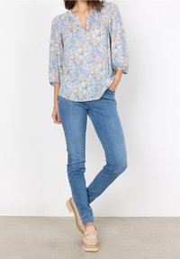 Blusa floreale azzurro chiaro con maniche a 3/4 e scollatura a V, abbinata a jeans blu slim-fit e mocassini beige con platform su un pavimento neutro.