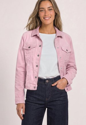 Jeune femme souriante portant une veste rose clair boutonnée, un t-shirt blanc et un jean bleu foncé, une main dans la poche, sur un fond uni.