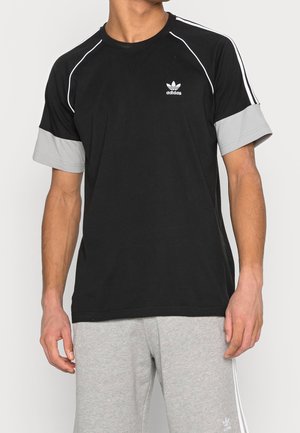 Mężczyzna w czarnej koszulce Adidas z jasnoszarymi mankietami na rękawach oraz w dopasowanych szarych spodniach dresowych Adidas.