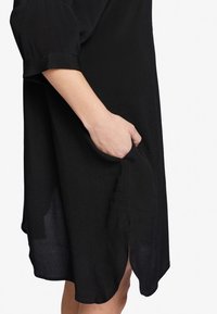 Svart klänning med en lös passform, tillverkad av lättviktsmaterial. Har en sidoficka och en sidoklyvning, med 3/4 ärmar och en mjuk yta.