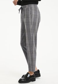 Pantalon à carreaux en gris, noir et marron. Doté d'une taille élastique avec cordon de serrage et de jambes fuselées. Porté avec des chaussures à plateforme noires.