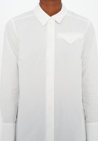 Homme portant une chemise blanche à manches longues boutonnée, avec un col pointu et une poche à rabat triangulaire sur la poitrine gauche.