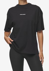 Person wearing a black oversized T-shirt with "PEGADOR" text on the chest and black leggings, hand pulling the shirt slightly to the side.