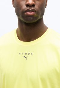 Κοντινό πλάνο ενός ατόμου που φοράει ανοιχτοκίτρινο πουκάμισο με το λογότυπο «HYROX» και ένα λογότυπο Puma τυπωμένα σε γκρι στο στήθος.