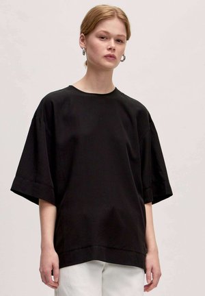 Black oversized short-sleeve top with a round neckline, smooth texture, and side slits, paired with light-colored pants.