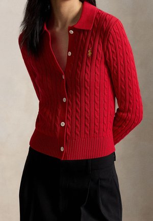 Frau trägt eine rot geknöpfte Zopfmusterstrickjacke mit Kragen und schwarze Hose, vor neutralem Hintergrund.