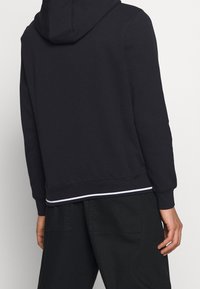 Marinblå hoodie med dragsko, ribbade ärmavslut och nederkant med en vit accentrand, tillverkad av mjukt, texturerat material.