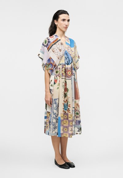 Femme aux cheveux foncés portant une robe patchwork ample mi-longue avec des motifs floraux et géométriques colorés, associée à des ballerines noires.