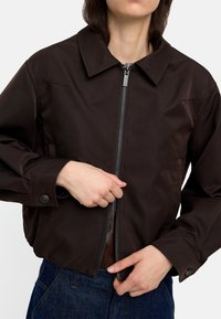 Brązowa krótka kurtka z wyprofilowanym kołnierzem, zamkiem błyskawicznym z przodu i zapięciem na guziki przy mankietach. Materiał wydaje się gładki z minimalnymi detalami przeszycia.