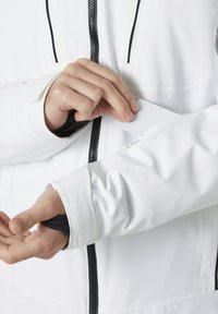 Bílá měkká bunda s černým zipem a akcenty. Levý manžet m�á malý otvor pro přístup k kartě, který zobrazuje podrobné šití.
