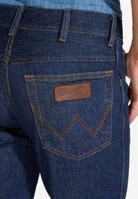 Jeans de mezclilla azul oscuro con una etiqueta de cuero Wrangler en el bolsillo trasero. Presenta costuras naranjas y un diseño distintivo de bolsillo en forma de chevrón.