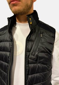 Gilet rembourré noir avec fermeture éclair à l'avant et poches. Présente un col haut et des accents texturés noirs sur les épaules. Porté par-dessus une chemise blanche.