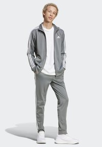 Sivá tepláková bunda s bielymi pruhmi na rukávoch, kombinovaná s zodpovedajúcimi nohavicami. Pod ňou biele tričko, nosené s bielymi športovými topánkami.