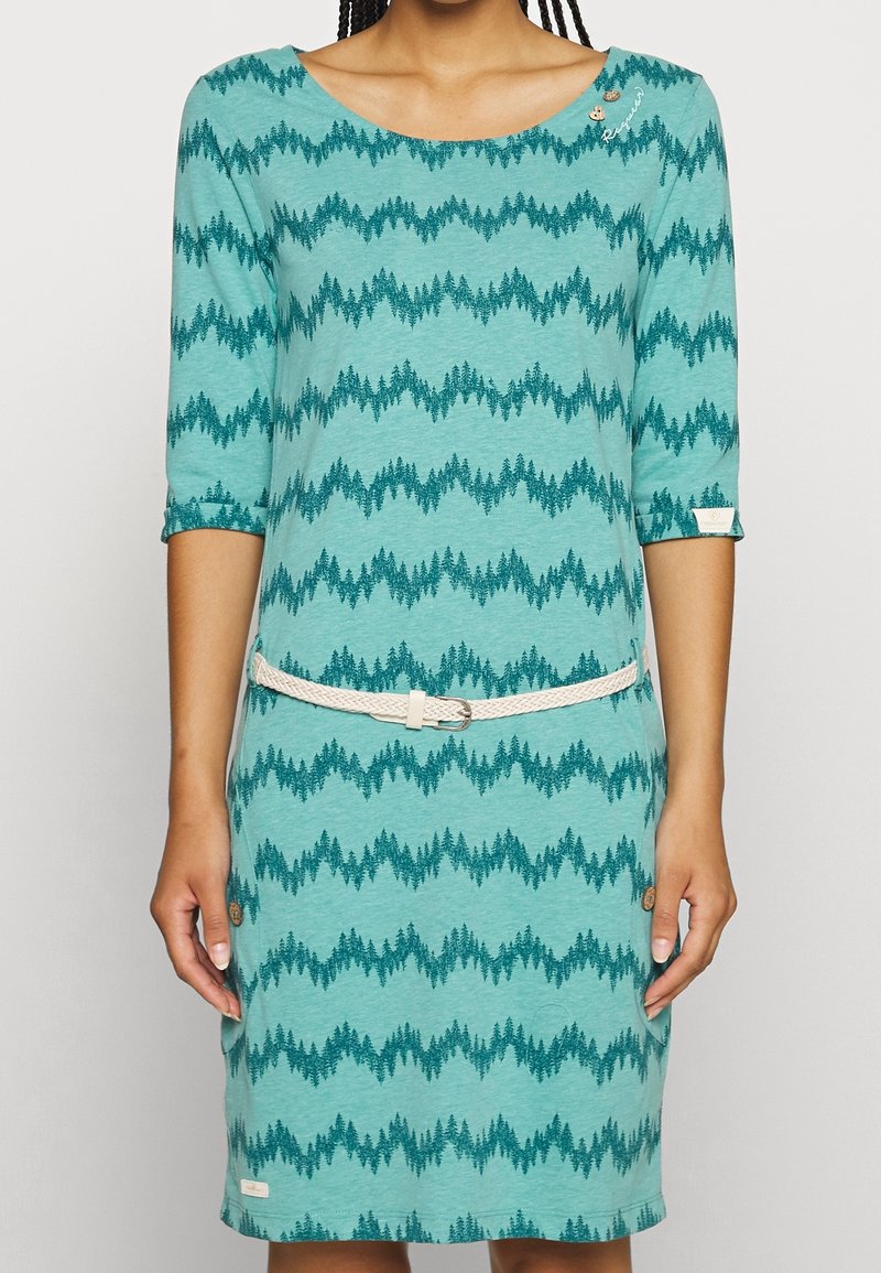 Turkusowa sukienka z wzorem fal w ciemnozielone drzewa, rękawy 3/4 i biały pleciony pasek w talii. Okrągły dekolt.