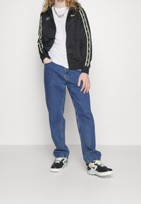 Černá bunda Nike s neonově zelenými prvky, bílé tričko, modré džíny a černé tenisky Nike se světle zelenými detaily.