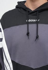 Čierna a fialová mikina s kapucňou, s bielym logom Adidas, sťahovacími šnúrkami a bielymi pruhmi na bokoch. Textúrovaná látka.