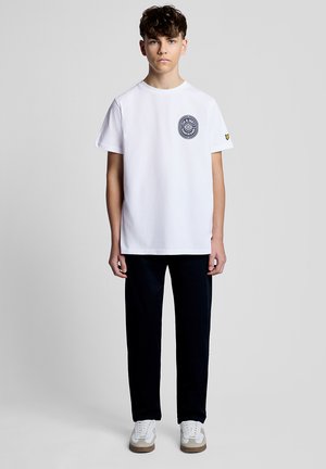 Teenagedreng iført en hvid t-shirt med grafik, sorte bukser og hvide sneakers, stående foran en ensfarvet lys grå baggrund.