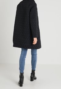 Sort quiltet frakke med løs silhuet, der har en høj krave og lange ærmer. Båret over blå jeans og sorte ankelstøvler.