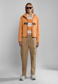 Oranje waterdichte jas met capuchon, twee voorzakken en logo-detail, gelaagd over een gestreept shirt, gecombineerd met beige cargobroek en sneakers.