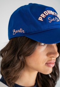 Căciulă de baseball din bumbac albastru, cu text brodat roșu și alb care spune "PROHIBIT" și "Social Club", având "Berlin" cusut în roz.