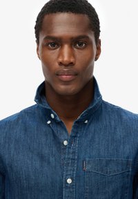 Camisa de denim en azul oscuro con cuello abotonado, botones blancos y un bolsillo en el pecho que presenta una etiqueta con logo. Tejido suave y texturizado.