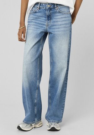 Personne portant un jean large bleu clair, des baskets blanches et une chemise blanche, debout devant un fond clair uni.